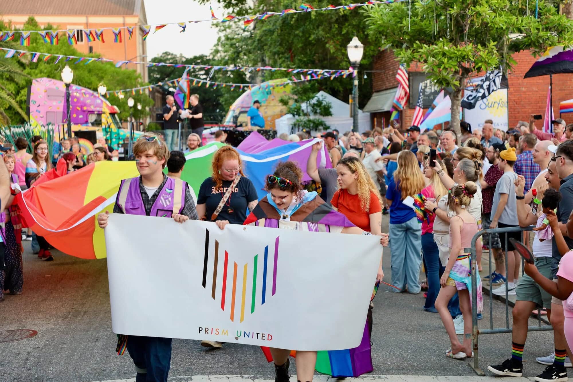 People march down a street during a Pride parade, holding a large Prism United banner, with rainbow flags and a crowd of onlookers in the background.