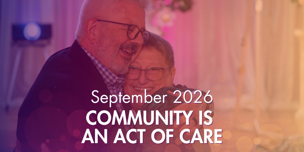 An older man and woman hug and smile in a warmly lit room. Text over the image reads, “September 2026. Community is an act of care.”.