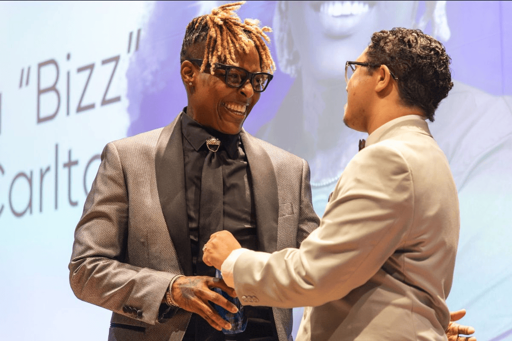 Two people in formal attire smiling and talking on a stage in front of a large screen displaying the name “Bizz” Carlton.