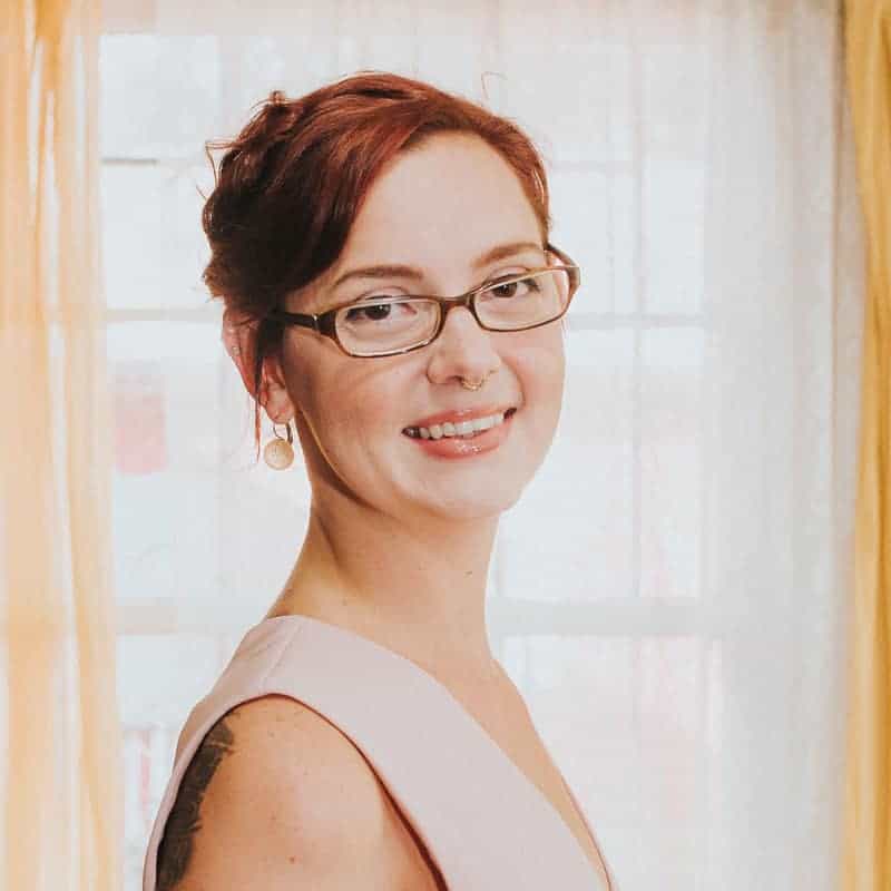 Woman with red hair and glasses smiling in front of a window with yellow curtains.