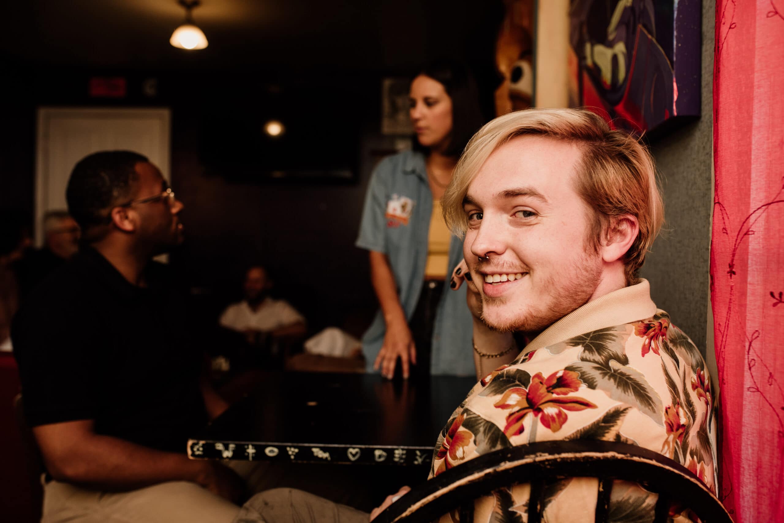 A young man in a patterned shirt smiles at the camera while seated in a dimly lit room with other people.
