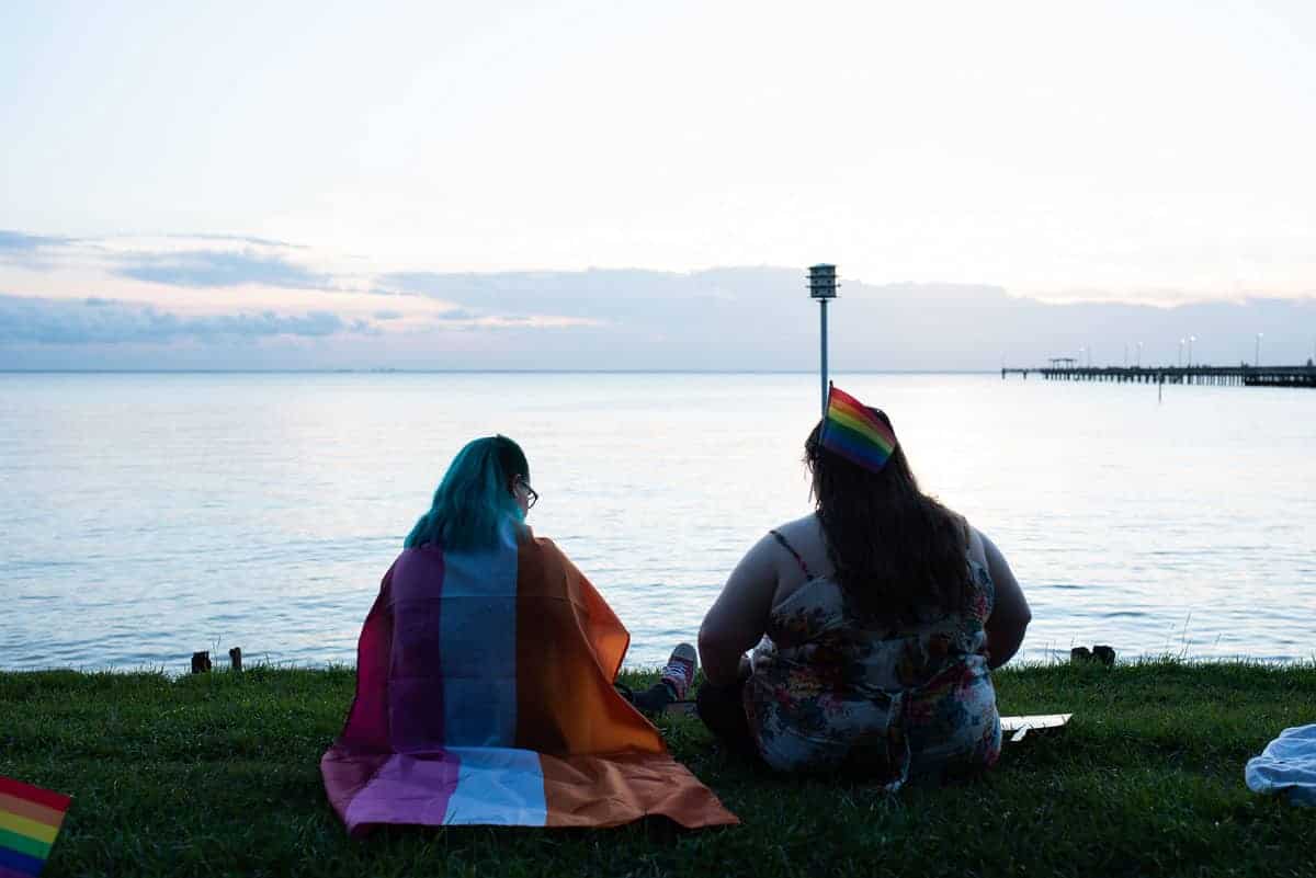 Two individuals with pride flags sitting by a lakeside at dusk.