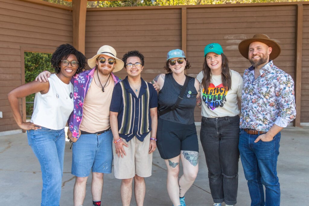 Six people stand close together outdoors, smiling at the camera. They wear casual summer clothing, hats, and sunglasses. A wooden building and trees are visible in the background.