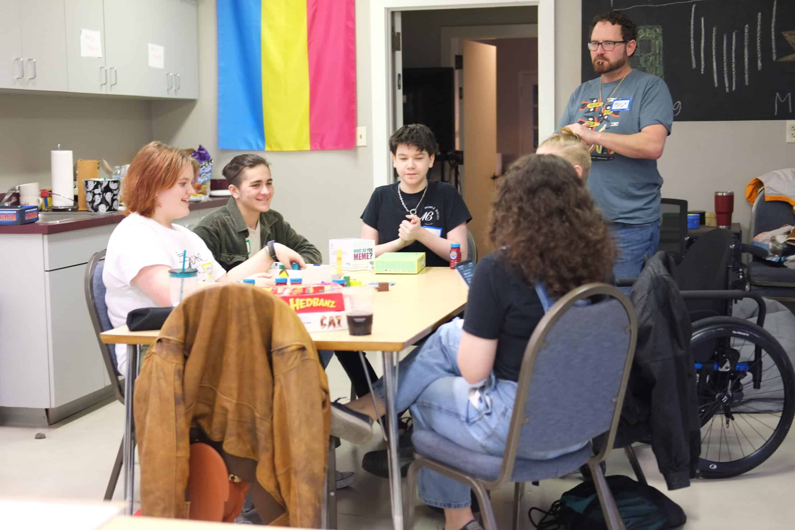 Group of individuals sitting around a table interacting, with games visible, in a room with a pride flag on the wall.